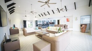 A spacious living room with multiple seating areas, ceiling fans, and a kitchen in the background.