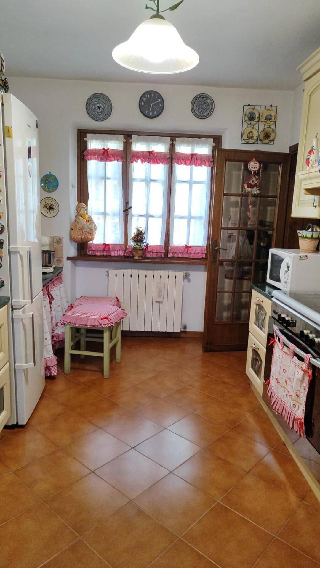 Una cocina con piso de baldosas, un taburete de madera, un refrigerador, un microondas y una ventana con cortinas.