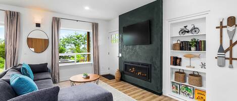 A modern living room with a grey sofa, a round mirror, a fireplace, and a bookshelf with various items.