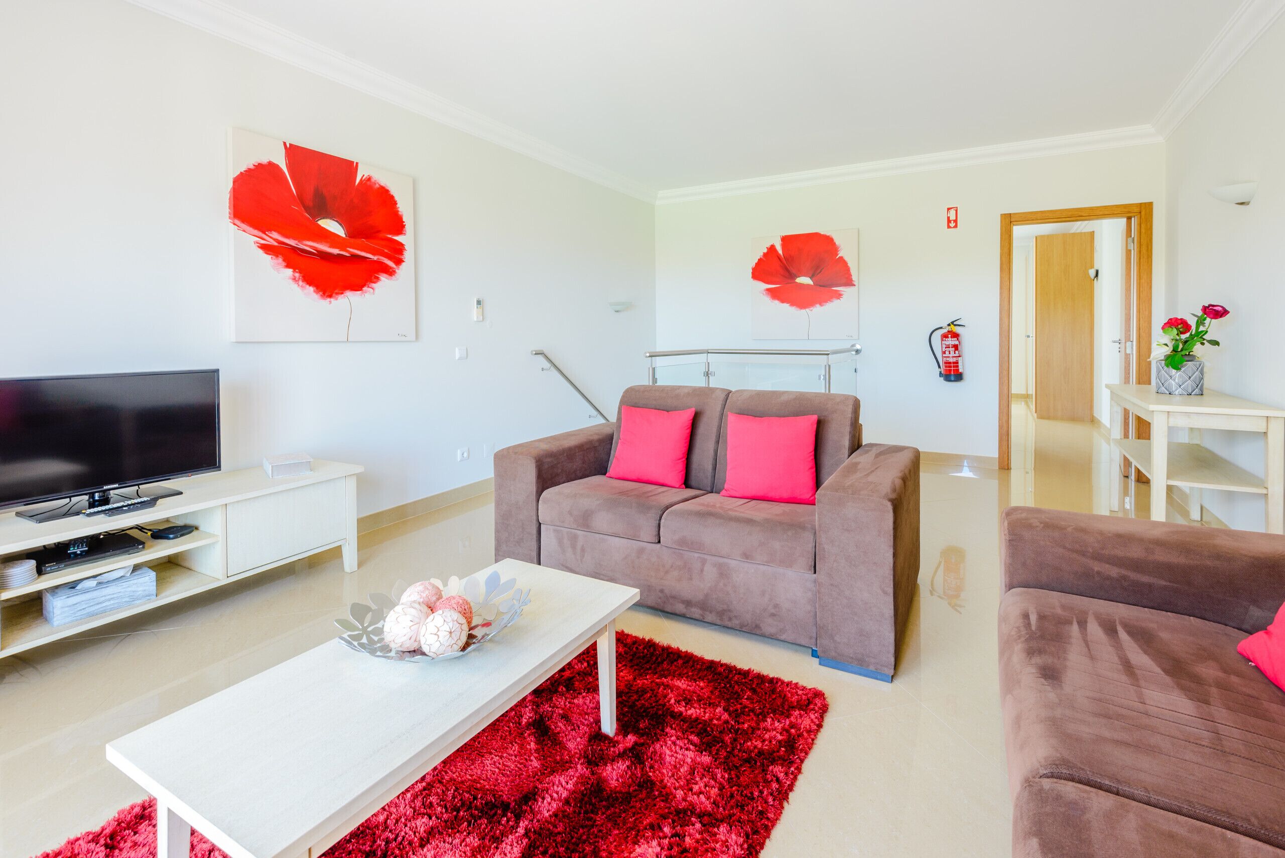 A modern living room with a flat-screen TV, a sofa with pink cushions, a coffee table with decorative items, and red pop art on the walls.