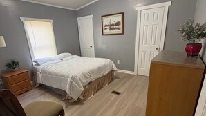 A bedroom with a bed, a chair, a dresser, a window with blinds, and a door.