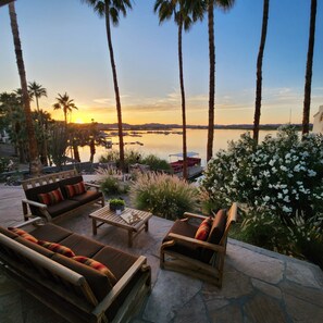 A patio with outdoor furniture, a view of the sunset over a body of water, and palm trees.