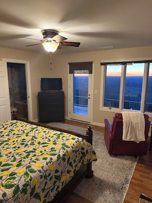 A bedroom with a bed, a ceiling fan, a dresser, a chair, and a balcony with a view.