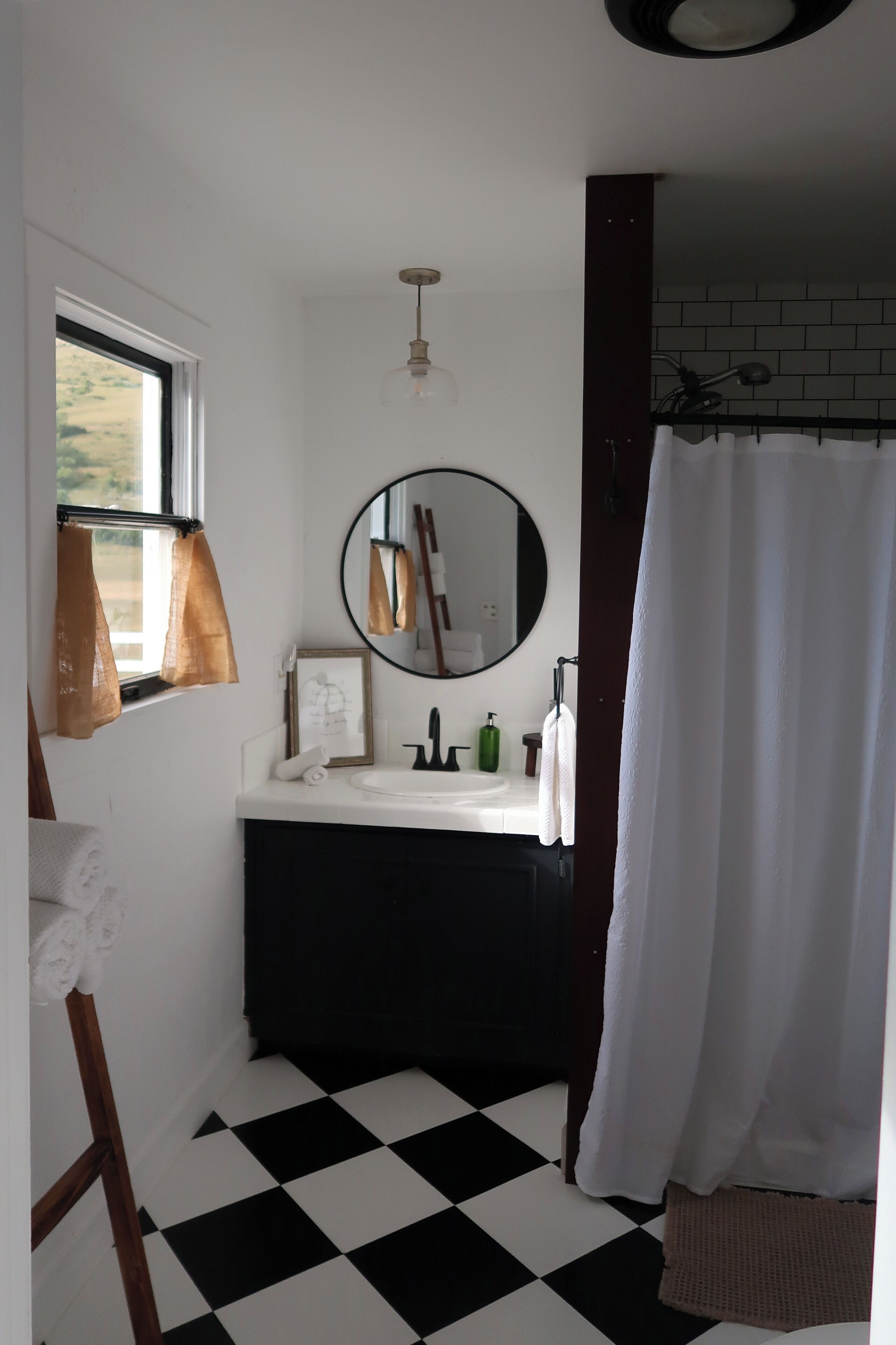 Un baño con piso a cuadros blanco y negro, un espejo redondo, un lavamanos blanco y una ventana con cortinas marrones.