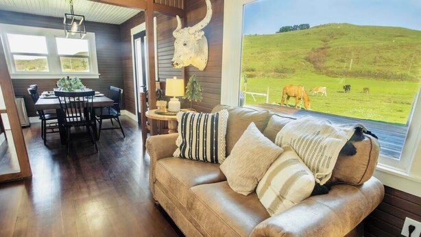 Un salon chaleureux avec un canapé en cuir, un coin repas avec une table et des chaises, et une grande fenêtre donnant sur un champ verdoyant et des animaux.