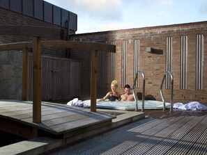 A rooftop pool area with two people relaxing in the water, wooden deck, and a brick building in the background.