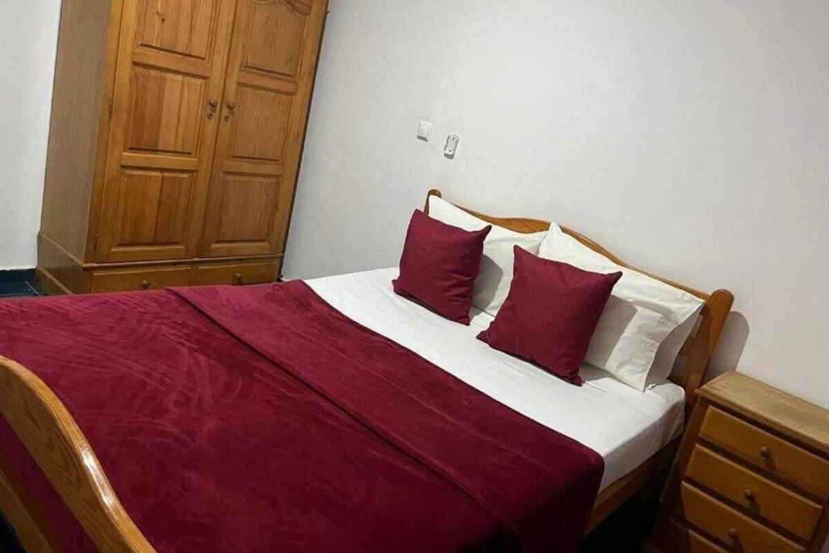A bedroom with a wooden bed, a red bedspread, two red pillows, a wooden nightstand, and a wooden wardrobe.