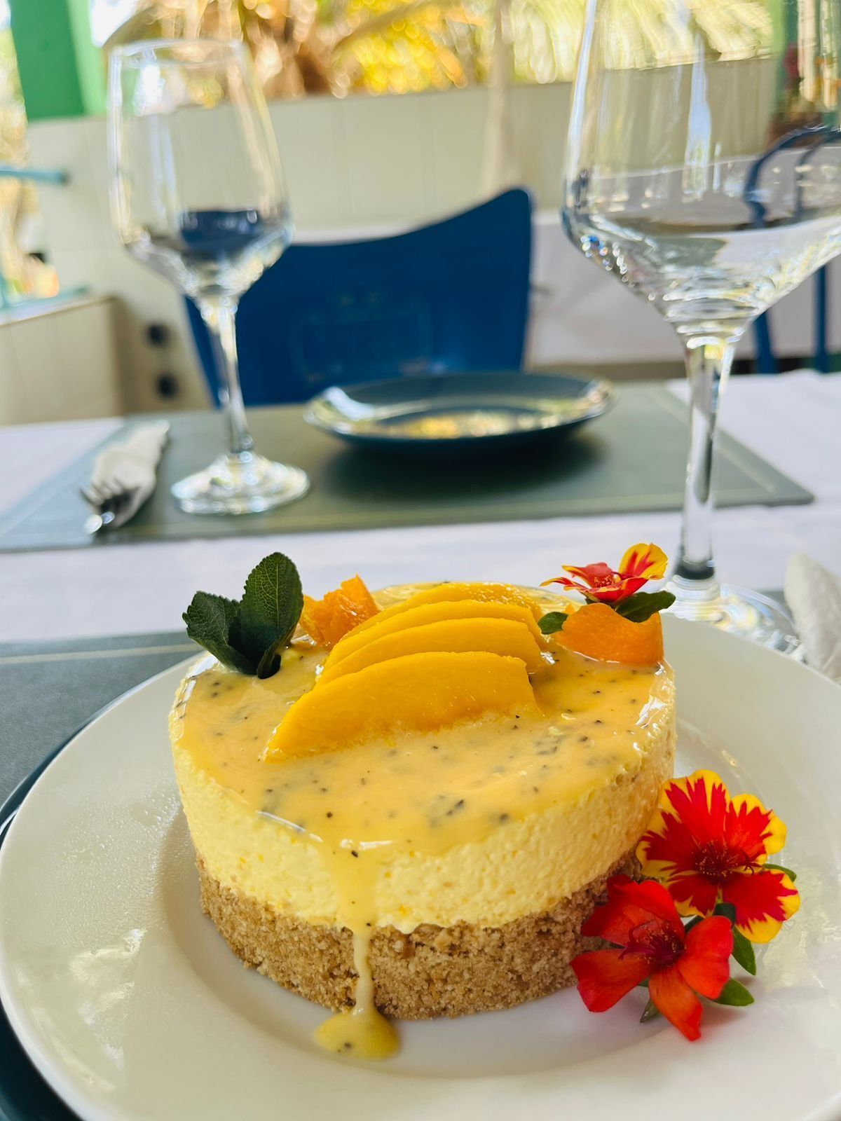 Un dessert présenté sur une assiette, composé d’une tranche de mangue et de quelques fleurs, accompagné de deux verres d’eau.