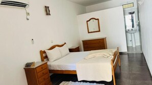 A bedroom with a bed, wooden nightstands, a dresser, a mirror, and a bathroom visible through an open door. - Hotel Village Penareia (Tarrafal)