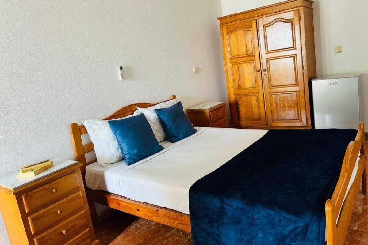 A bedroom with a bed, wooden wardrobe, nightstand, and a small white appliance.
