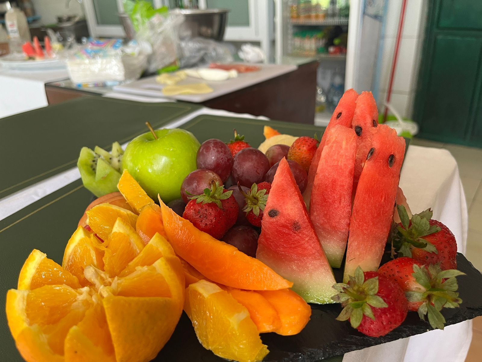 A fruit platter with sliced watermelon, strawberries, grapes, kiwi, and orange segments on a black slate plate.