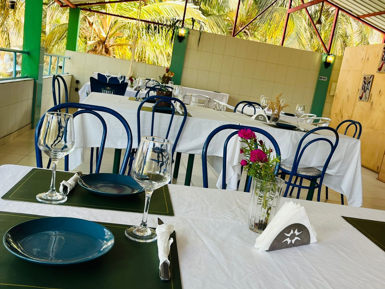 Un restaurant où les tables sont dressées pour le service, avec des chaises bleues, des nappes blanches et un décor tropical.