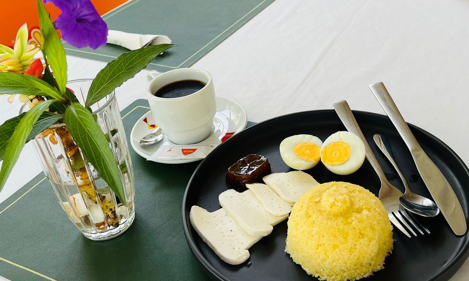 Un repas composé d’une assiette de riz jaune, d’œufs durs et de tranches de pain blanc, accompagné d’une tasse de café et d’un verre d’eau.