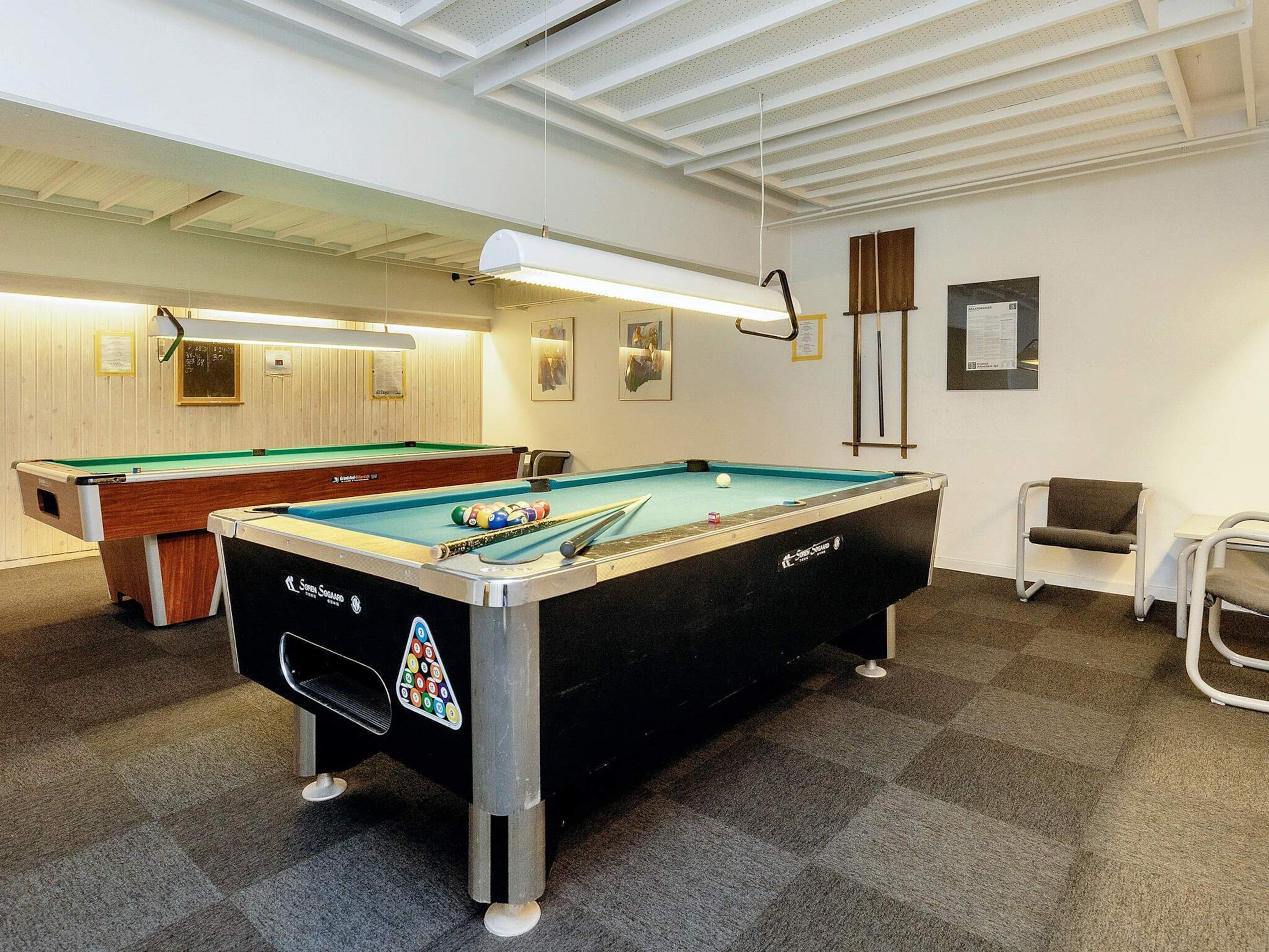 A pool table with a black tablecloth and a pool cue, a second pool table in the background, and a chair to the right.