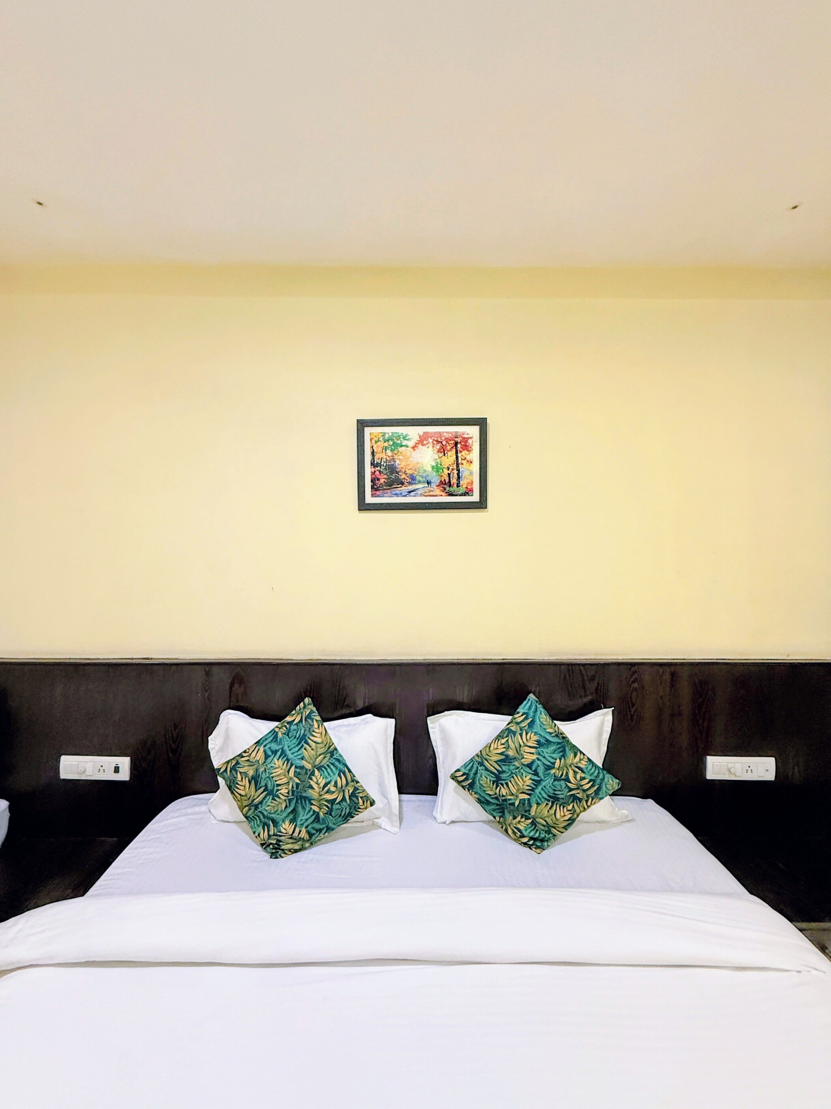 A hotel room with a bed, two decorative pillows, a wall-mounted painting, and a dark headboard.