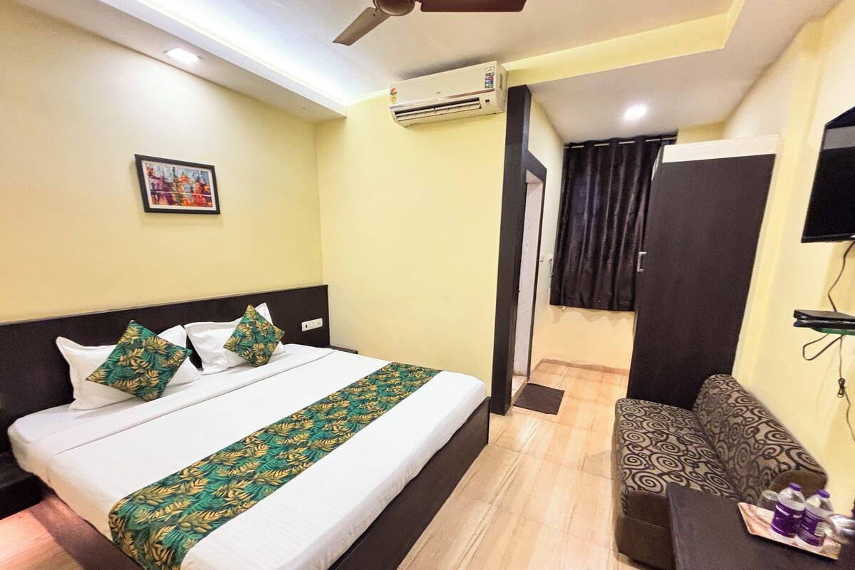 A hotel room with a bed, a sofa, an air conditioning unit, a television, and a door leading to another room.