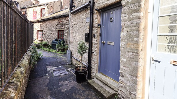 A narrow alleyway with stone buildings, a blue door, and a metal grate on the ground. - Olive Cottage (Kirkby Stephen)
