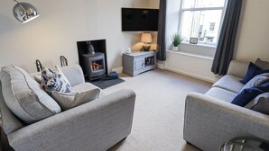 A modern living room with grey sofas, a flat-screen TV, a fireplace, and a window with curtains. - Olive Cottage (Kirkby Stephen)