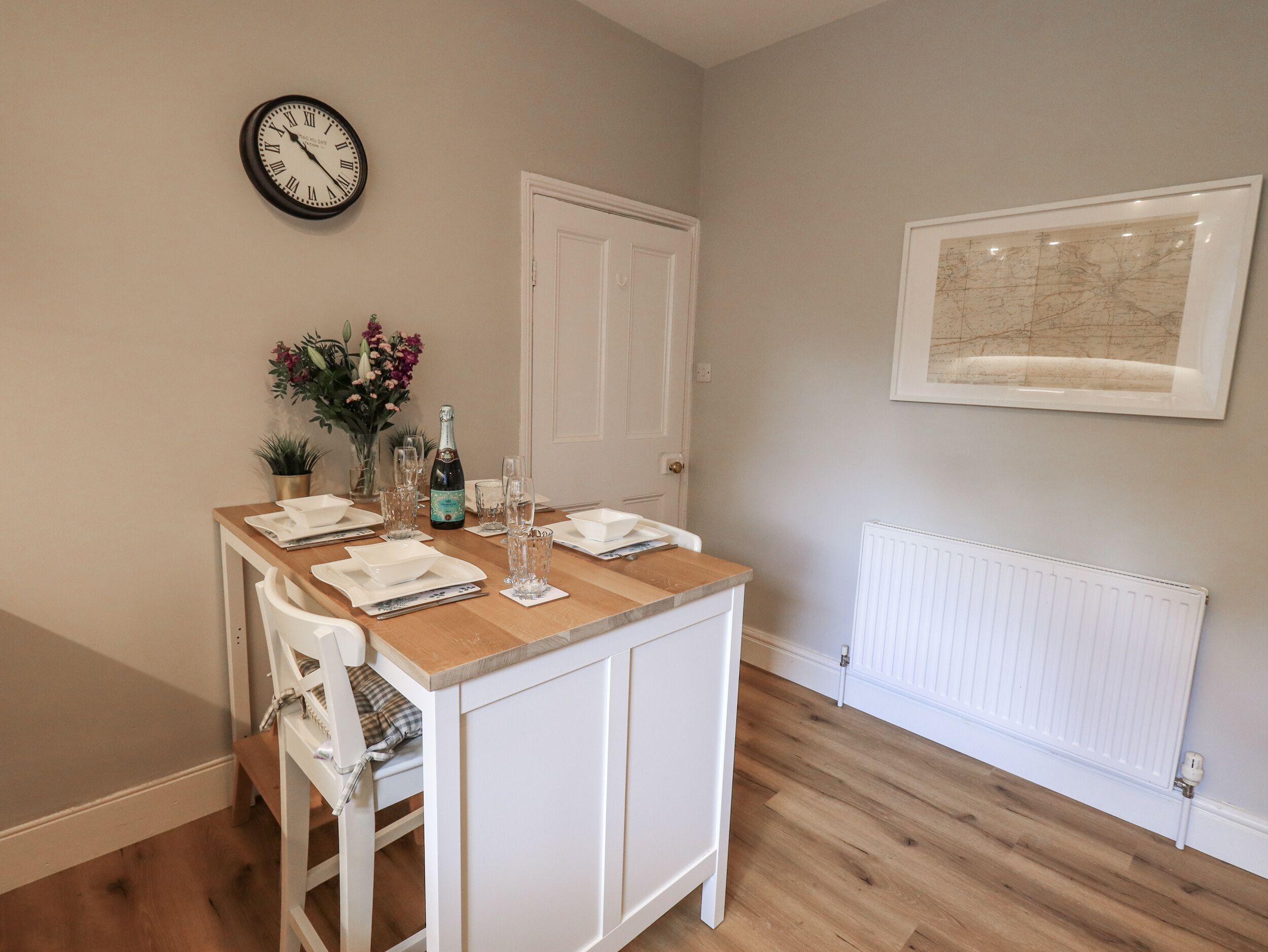 Un comedor con una mesa de madera preparada para cuatro personas, un reloj en la pared y un cuadro sobre un radiador.