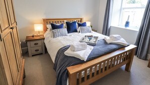 A neatly made bed with a wooden headboard, a bedside table with a lamp, and a window with curtains. - Olive Cottage (Kirkby Stephen)