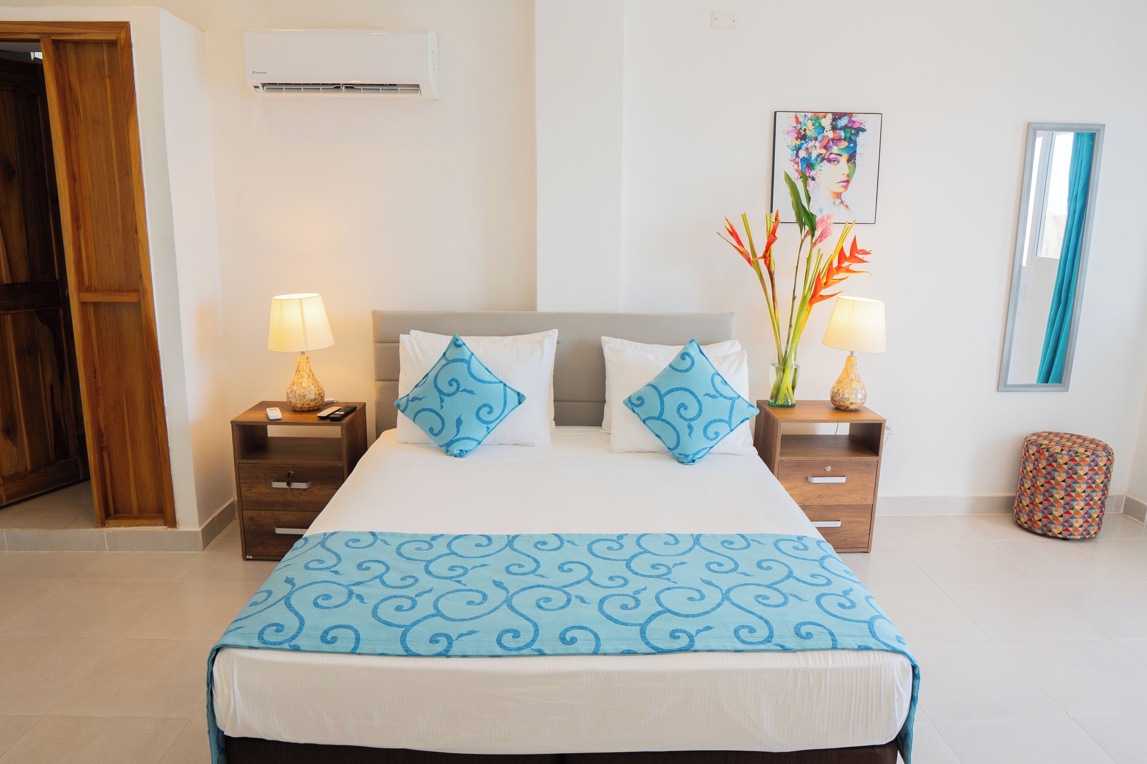A neatly arranged hotel room with a bed, two bedside tables with lamps, a small stool, a mirror, and a wall-mounted air conditioner.