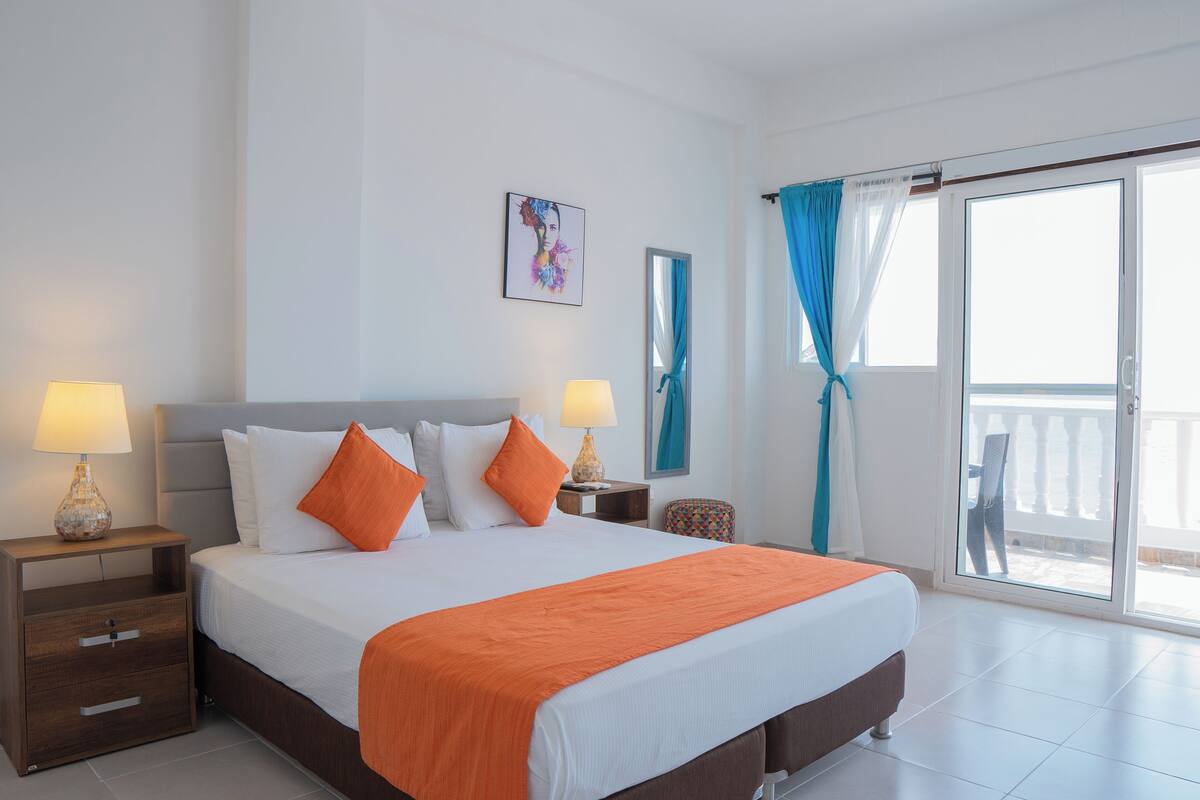 Una habitación de hotel con una cama grande, dos mesitas de noche con lámparas, un ventanal con cortinas y un balcón.