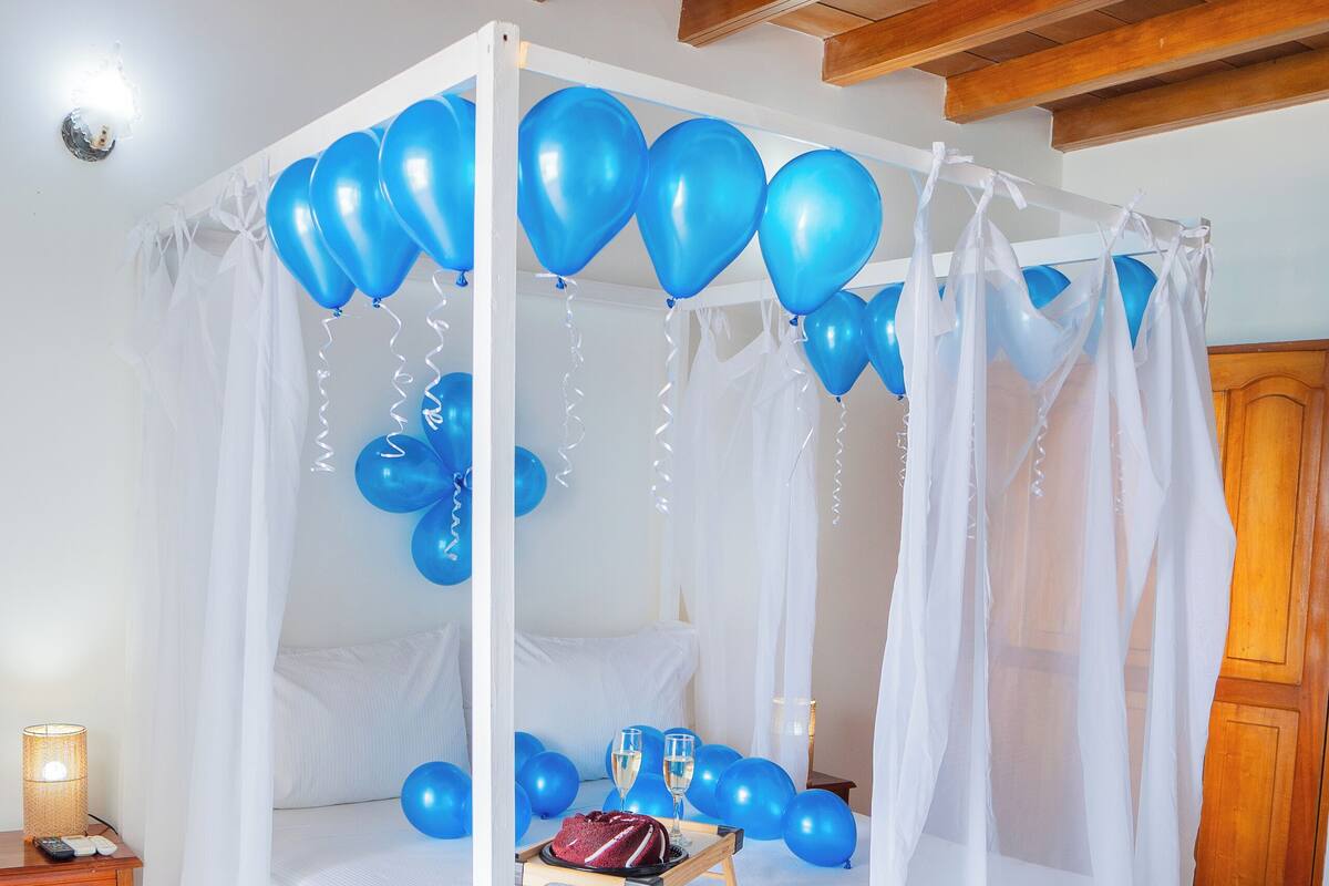 Una cama con dosel, rodeada de globos azules, una mesita de noche con una lámpara y suelo de madera.