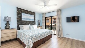 Ein Schlafzimmer mit Bett, Deckenventilator, Fernseher, Nachttisch, Lampe, einem Fenster mit Vorhängen und Blick auf den Balkon.