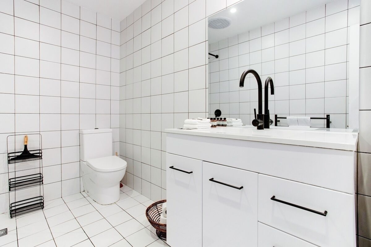 Ein modernes Badezimmer mit weißen Fliesen, einer weißen Toilette, einem schwarzen Wasserhahn und weißen Schränken.