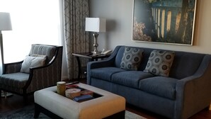 Un salon moderne avec un canapé bleu, un pouf assorti, un fauteuil à motifs, une table d’appoint avec une lampe et un tableau au mur.