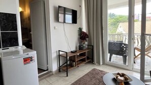 Un salon moderne doté d’un téléviseur à écran plat fixé au mur, d’un petit meuble en bois pour la télévision, d’un réfrigérateur et d’un balcon aménagé avec du mobilier d’extérieur.