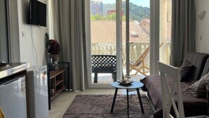 Un salon moderne comprenant un canapé, une petite table ronde, un téléviseur fixé au mur et une porte-fenêtre coulissante donnant sur un balcon avec vue sur les bâtiments et les collines.