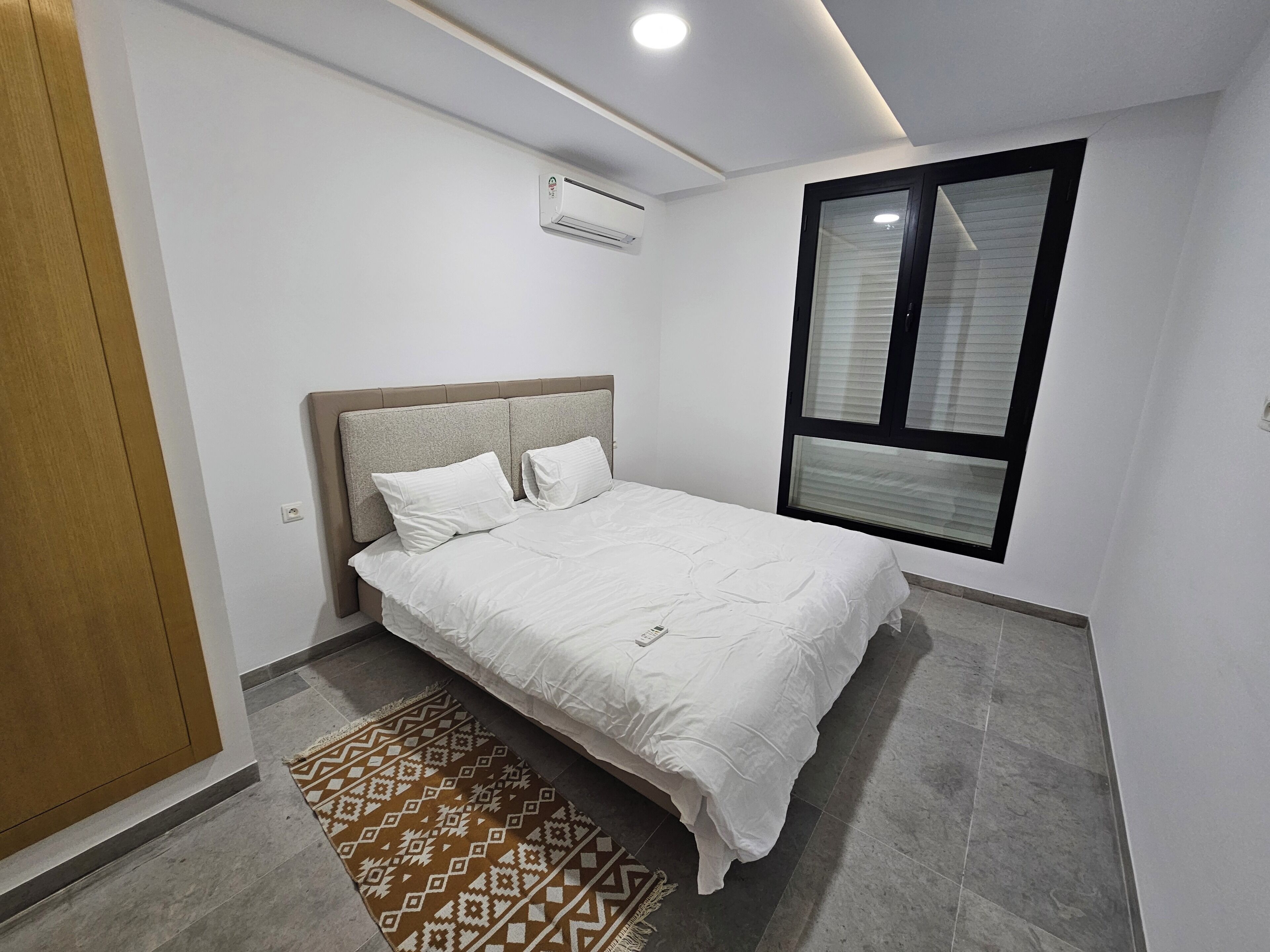 A bedroom with a bed, a window with blinds, an air conditioning unit, and a patterned rug.