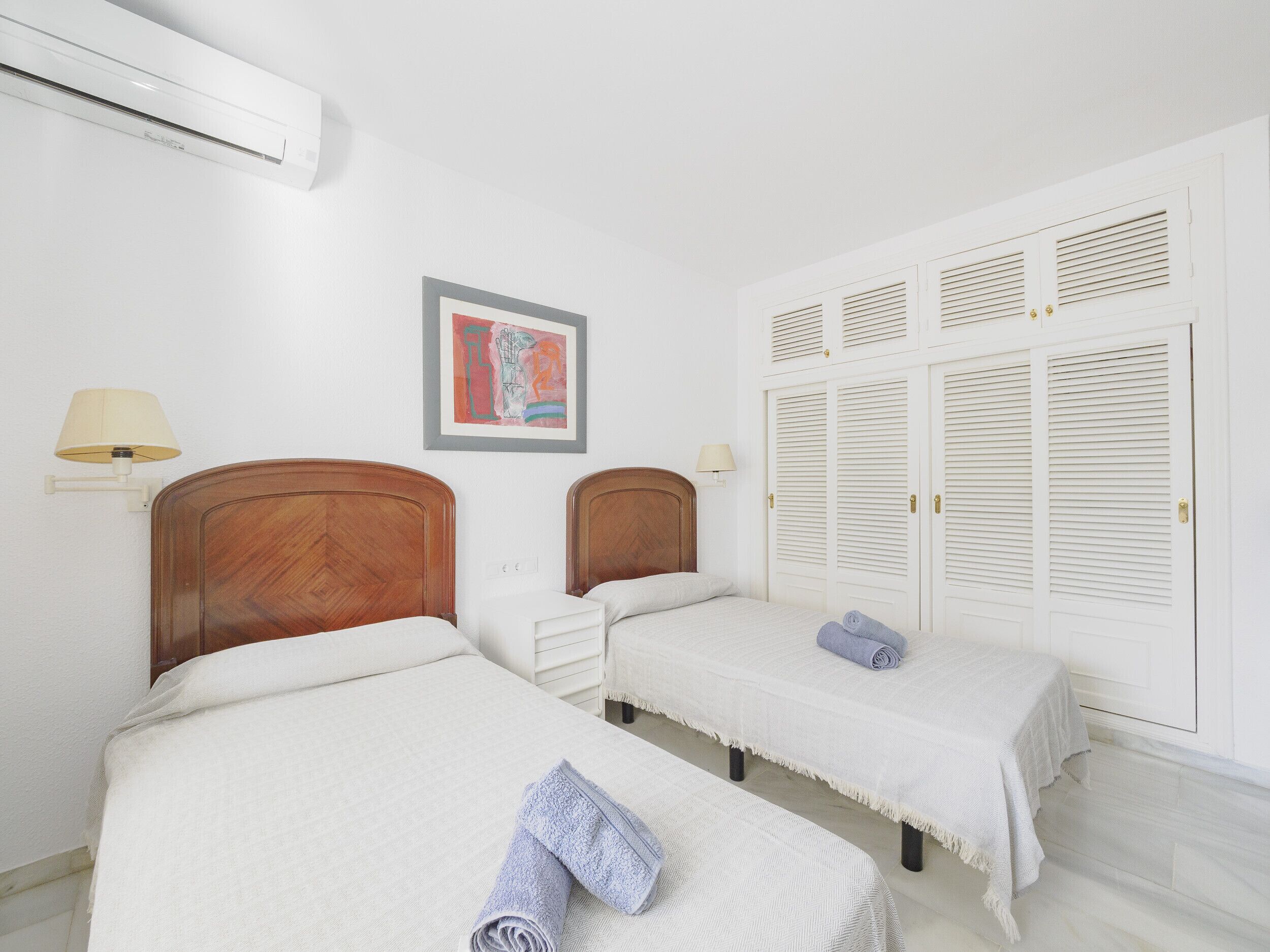 Une chambre d’hôtel avec deux lits, un climatiseur fixé au mur, un tableau encadré et un ensemble d’armoires blanches à persiennes.