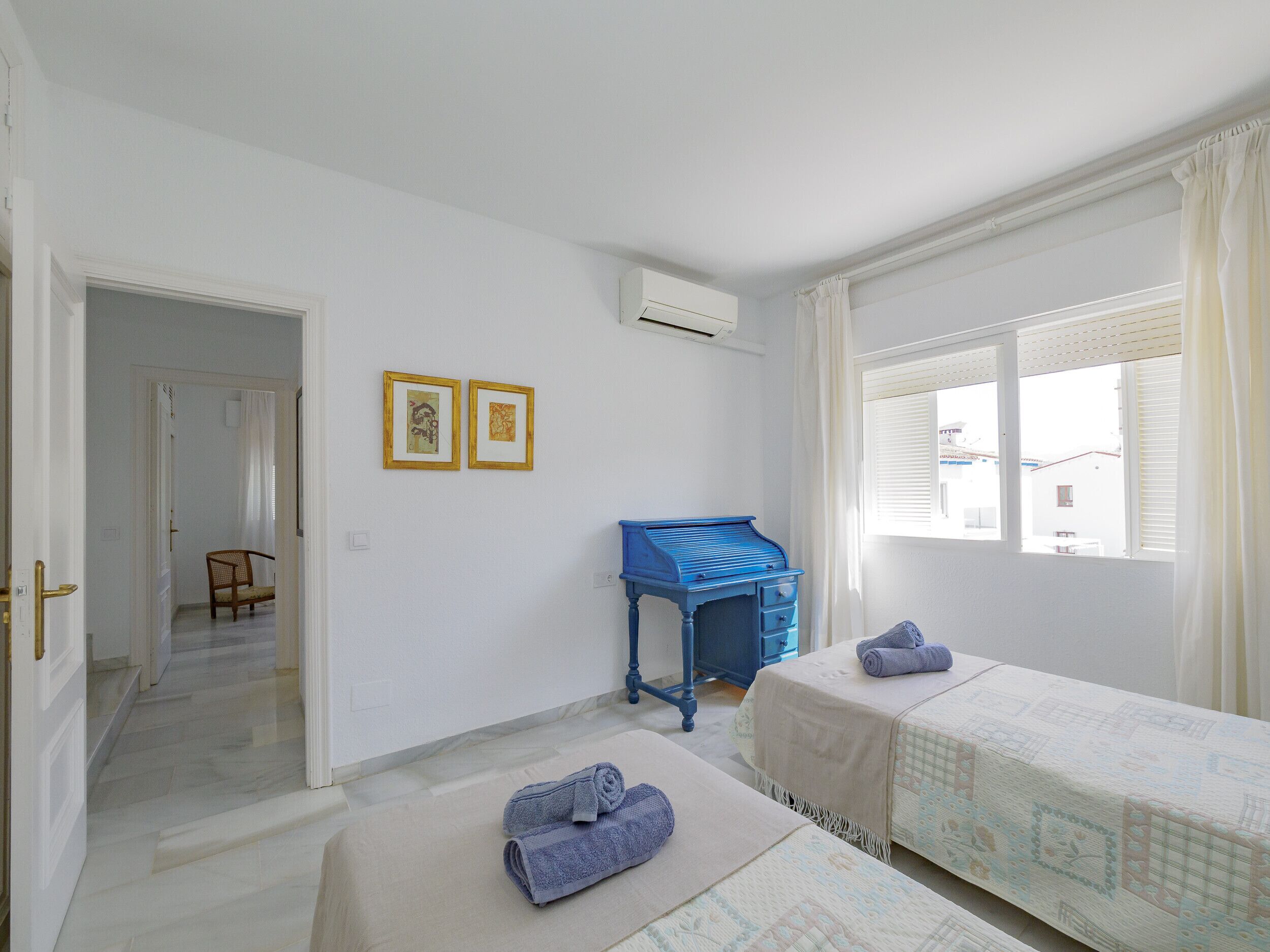 A hotel room with two beds, a desk, and a chair. There are two framed pictures on the wall.