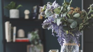 A dining table with a vase of flowers, a glass of orange juice, and a bowl of fruit.