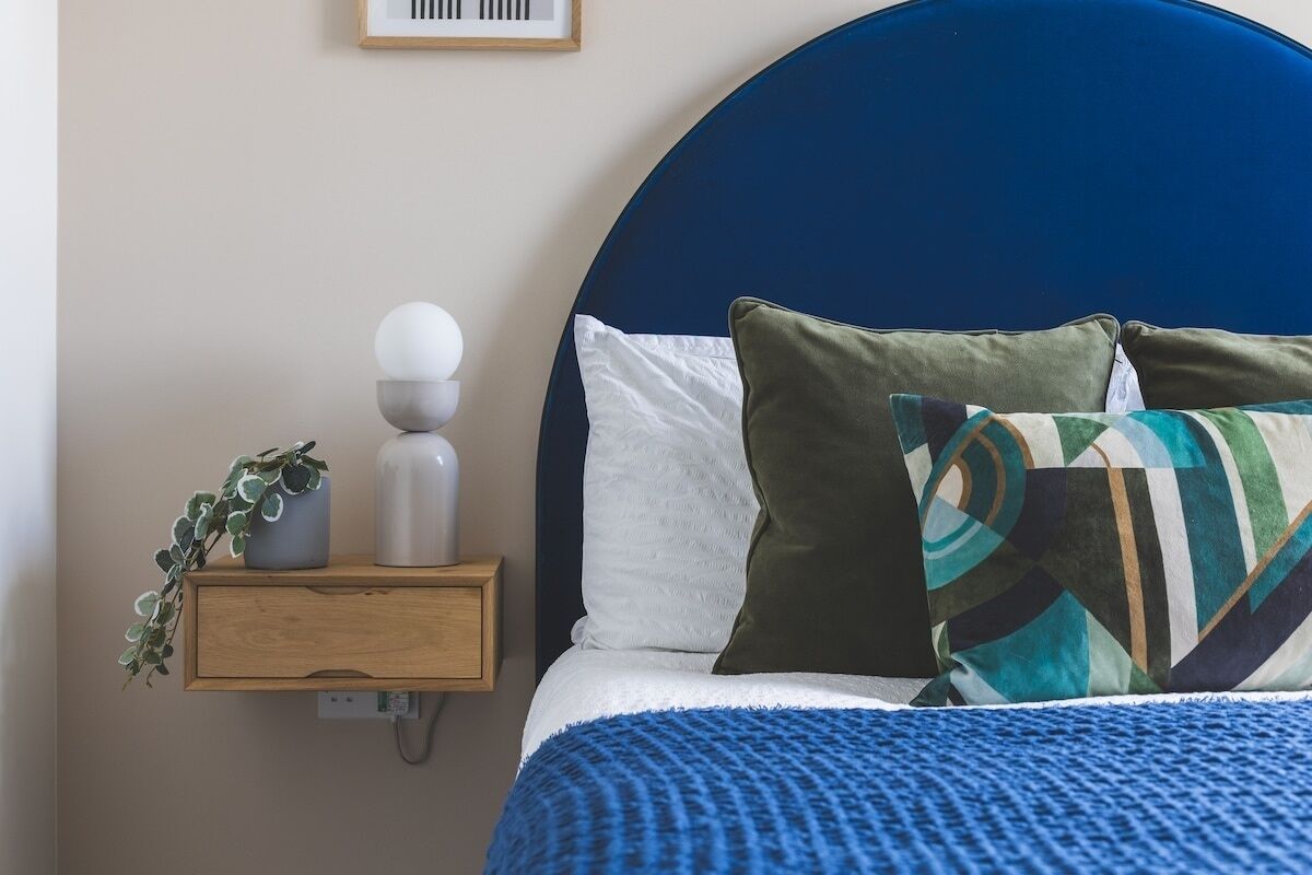 Una cama bien hecha con cabecera azul, una mesita de noche con una planta y un cuadro colgado en la pared.