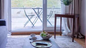Een gezellige woonkamer met een bank, een bijzettafeltje met kopjes en een schaal fruit, een klein houten tafeltje, en uitzicht op een balkon met tuinmeubilair.