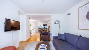 Een moderne woonkamer met een flatscreen-televisie, een bank, een salontafel met een afstandsbediening en een boek, en een eethoek op de achtergrond.