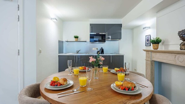 Een eetruimte met een ronde houten tafel gedekt voor het ontbijt, een keuken op de achtergrond en een open haard met decoratieve objecten.