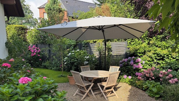A patio with a round wooden table and chairs, a large umbrella, and a garden with various plants and flowers.