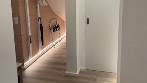 A hallway with wooden flooring, white walls, and a white door at the end. There is a coat rack with a black umbrella and a white coat hanging on it.