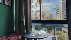 A room with a view of a cityscape, a table with two glasses, a hanging light fixture, and a bed with patterned bedding. - Vert House (Joao Pessoa)
