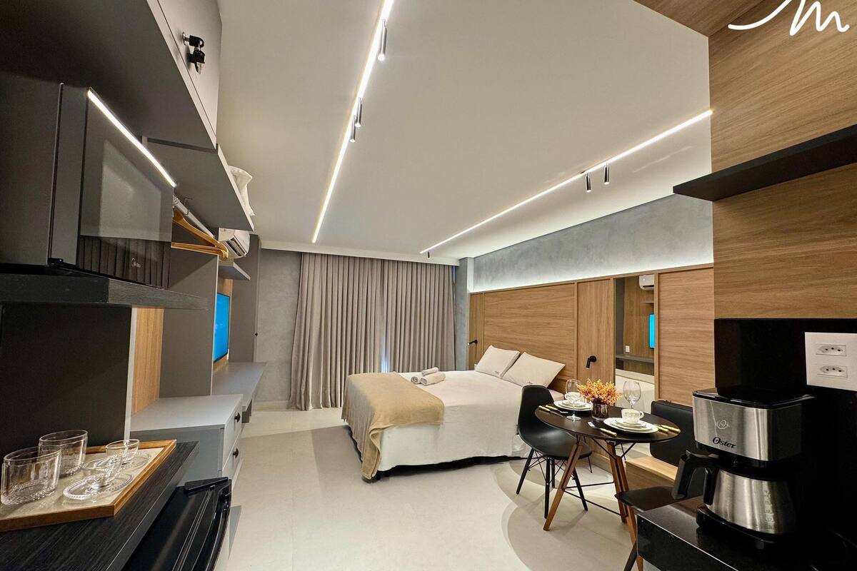 A modern hotel room with a bed, a dining area, a coffee machine, and a TV.