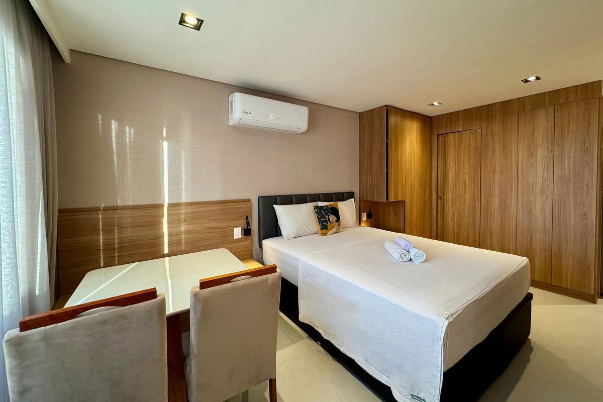 A hotel room with a bed, a desk, and a chair. There is an air conditioning unit on the wall.