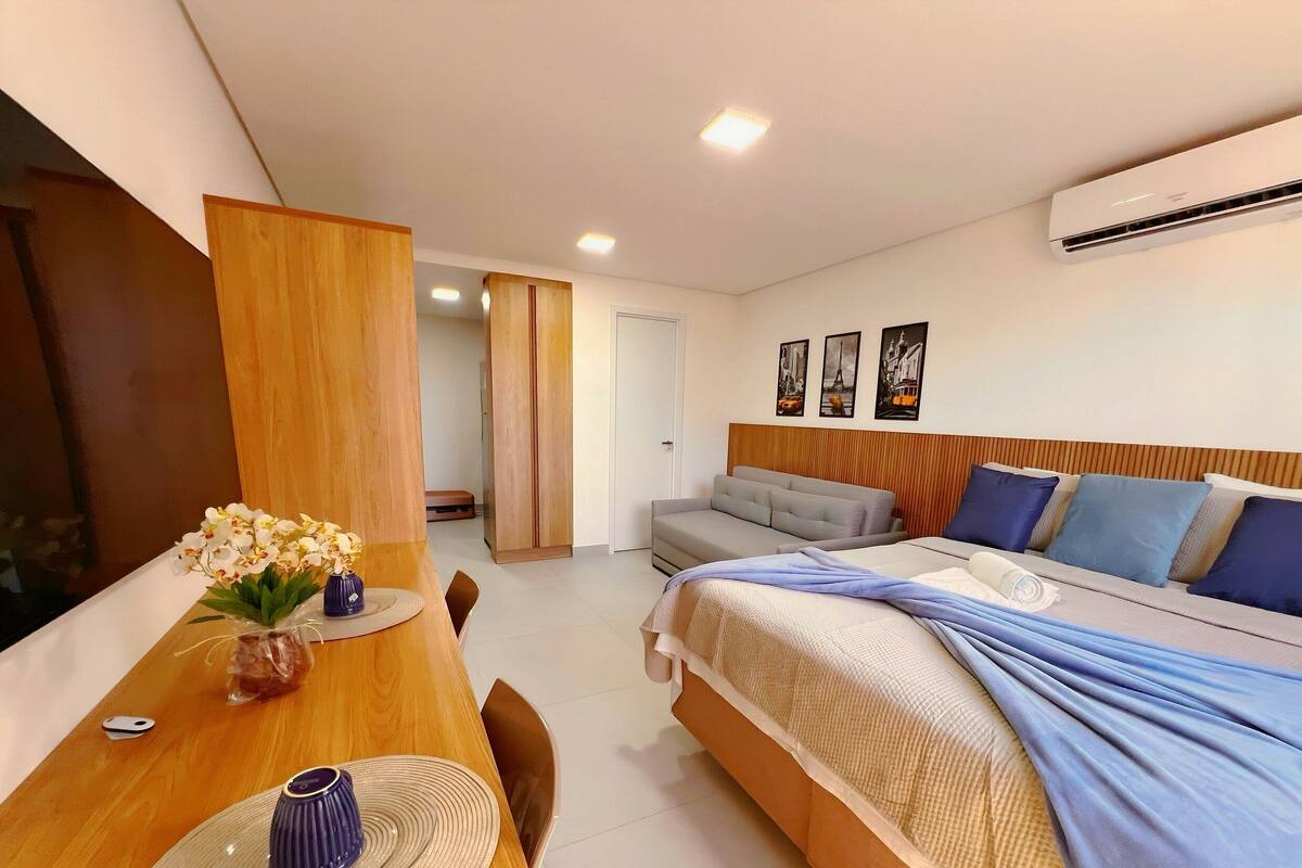 A modern hotel room with a bed, a sofa, a dining table with chairs, and an air conditioning unit.