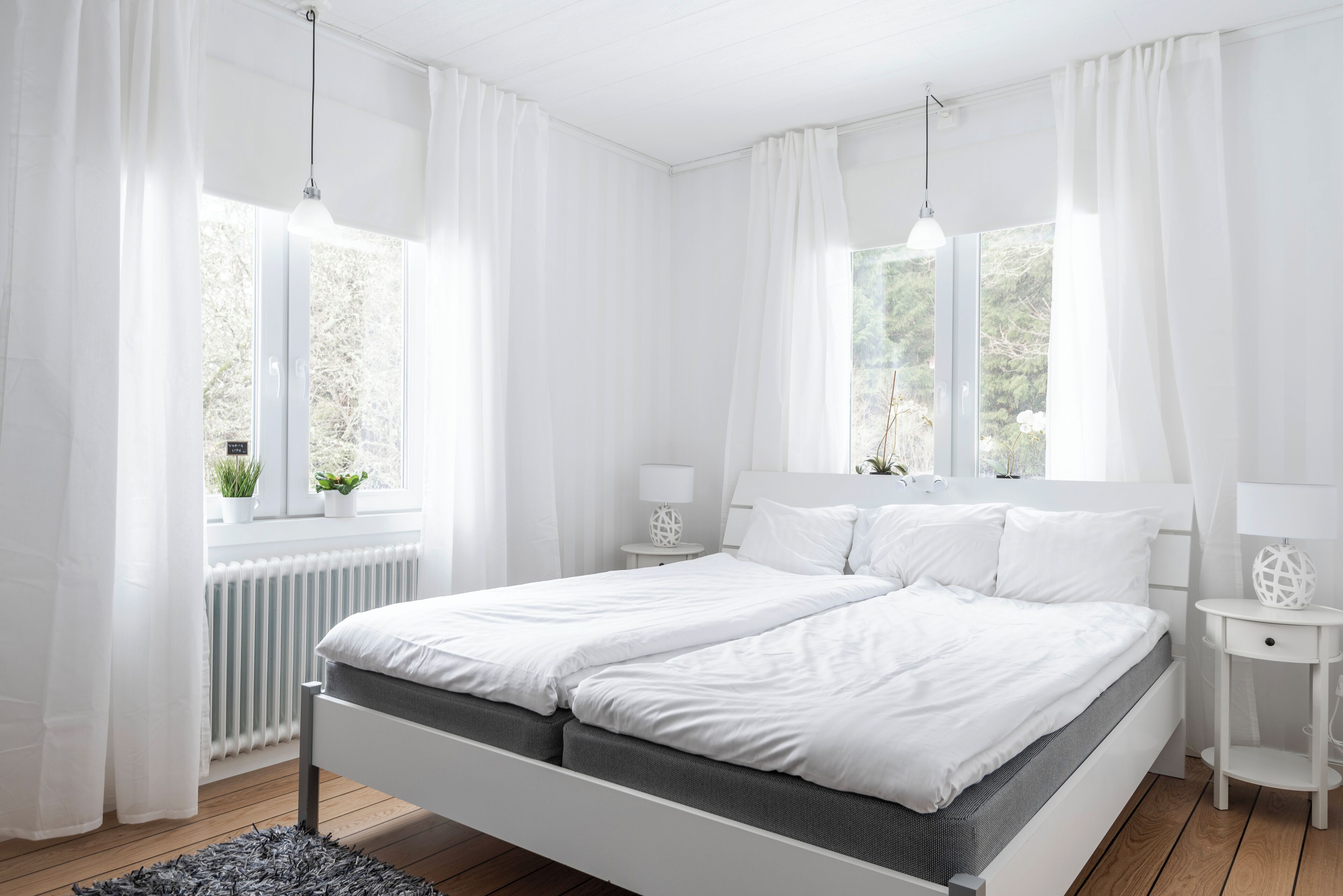 A modern bedroom with a double bed, white curtains, bedside lamps, and a view of greenery outside.