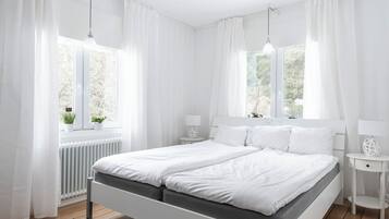 A modern bedroom with a double bed, white curtains, bedside lamps, and a view of greenery outside.