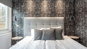 Camera da letto con testiera trapuntata, un letto grande con biancheria grigia, due comodini con lampade e una finestra che offre luce naturale.