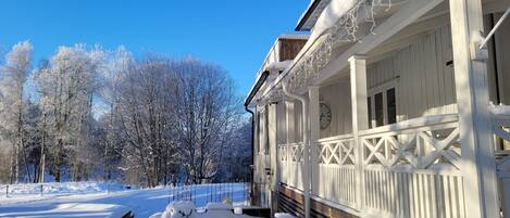 Un espacio exterior cubierto de nieve con un banco de madera y una mesa nevada, un edificio blanco con balcón, y una cerca al fondo.